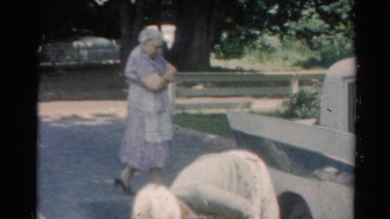 Woman standing near a white station wagon while another adult bends in the foreground.