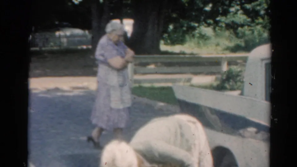 1964 Elkhart, Illinois home-movie frame showing a woman near a white station wagon in a shaded suburban yard.
