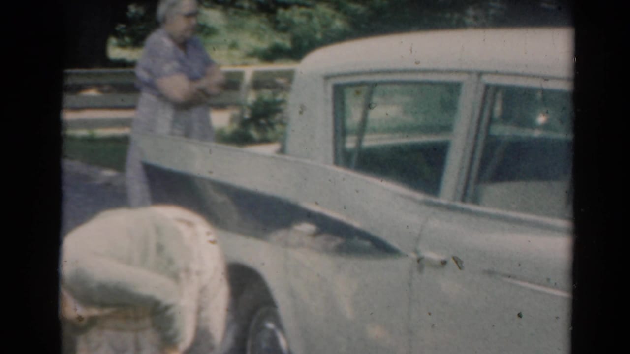 White station wagon beside a yard, with an adult standing near the rear area.