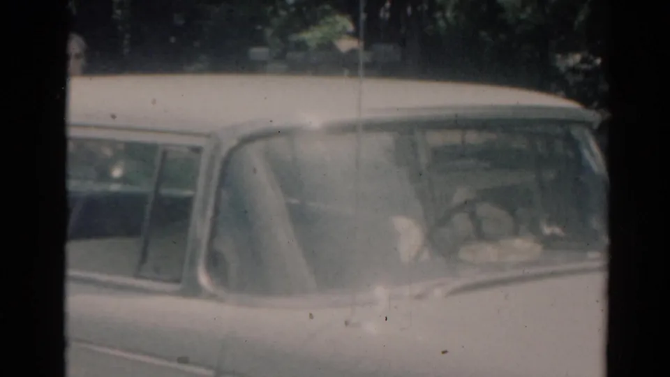 1964 Elkhart home-movie frame showing the rear of a white station wagon under trees near a residential yard.