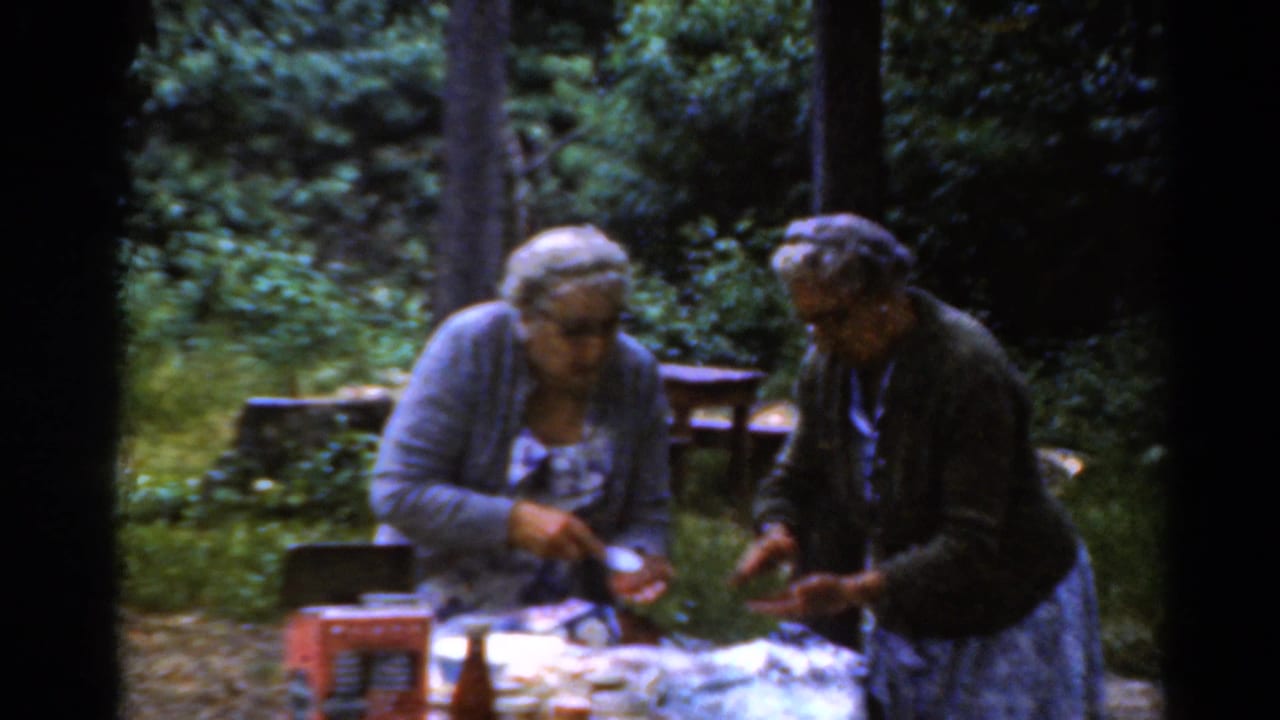 Two older women stand at a picnic table preparing food in a wooded area.
