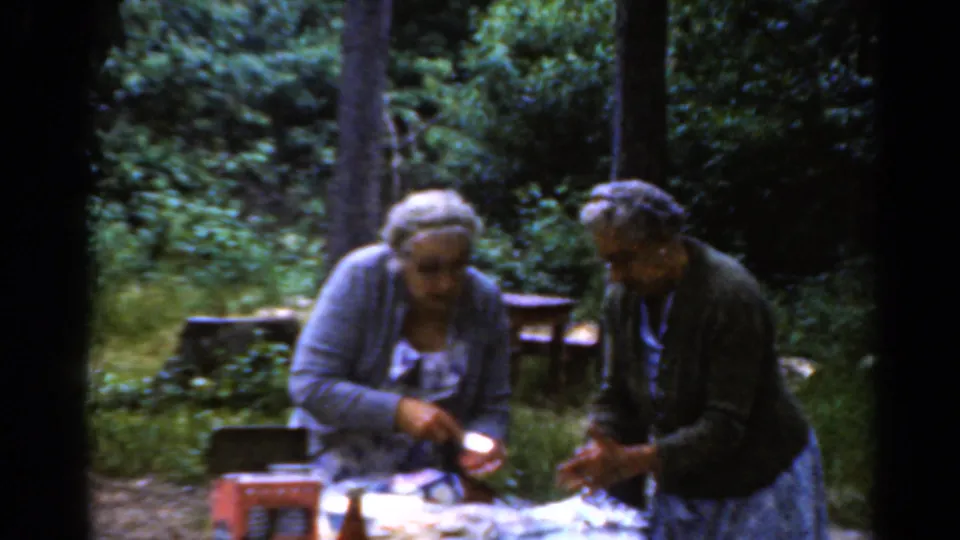 In the 1964 campground scene, two older women lean over a picnic table while handling food and small containers.