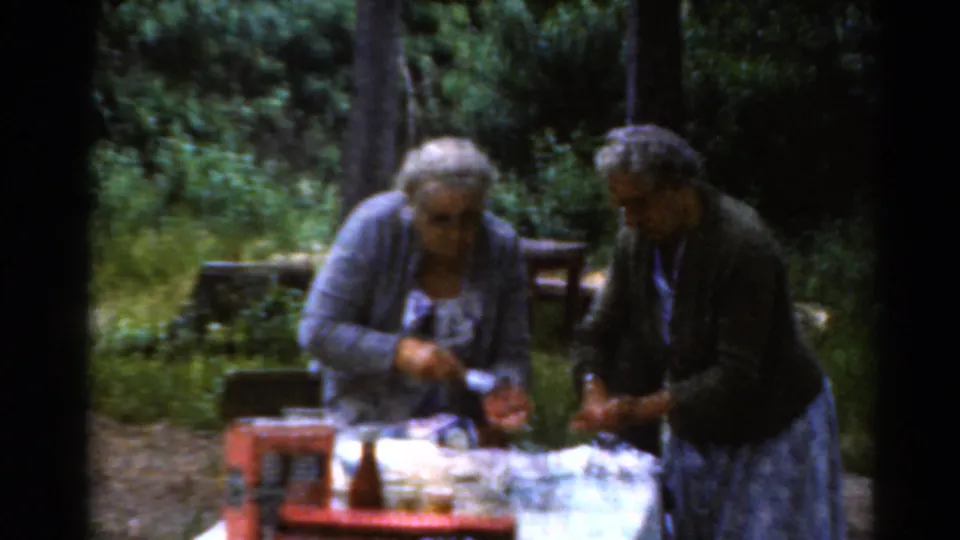Two older women in a wooded Elkhart, Illinois campground continue handling food at a picnic table in soft 8mm color footage.