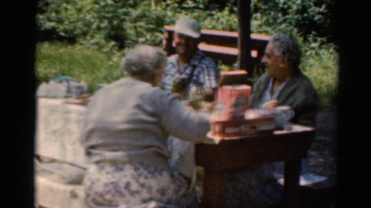 Three adults sit around a picnic table outdoors with food and drinks.