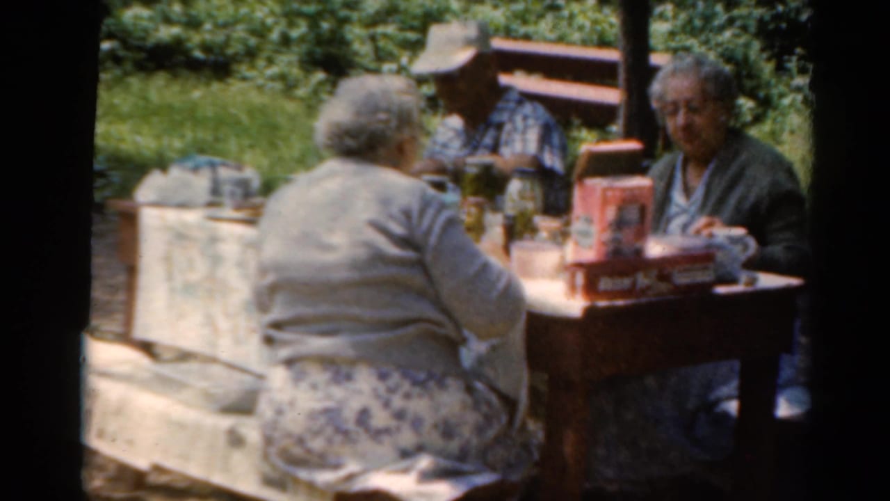 Adults at a wooden picnic table continue a casual lunch in 1964 Elkhart, Illinois home-movie footage.