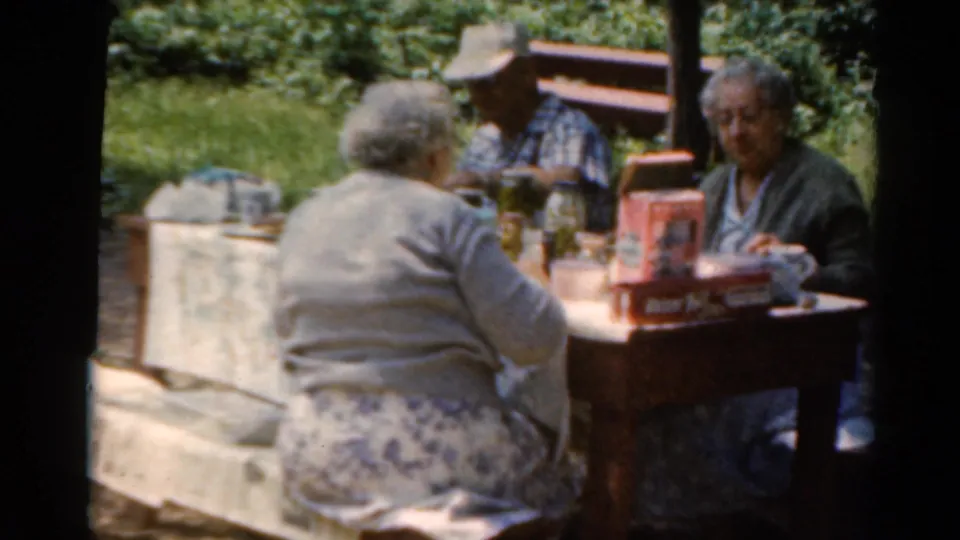 Adults at a wooden picnic table continue a casual lunch in 1964 Elkhart, Illinois home-movie footage.