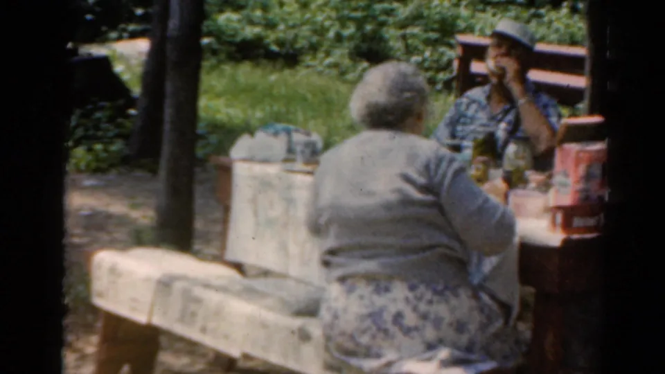 The handheld 8mm frame shows adults at a park picnic table, with one person seen from behind and others facing the camera.
