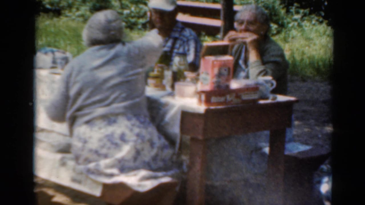 In a wooded Elkhart, Illinois picnic setting, older adults sit at a table with packaged food, bottles, and cups in 1964 home-movie footage.