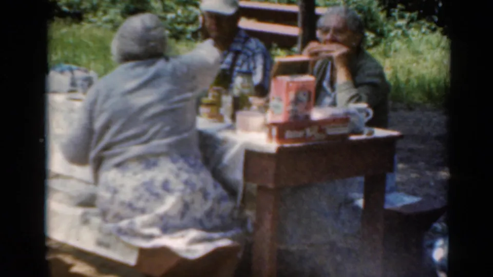 In a wooded Elkhart, Illinois picnic setting, older adults sit at a table with packaged food, bottles, and cups in 1964 home-movie footage.