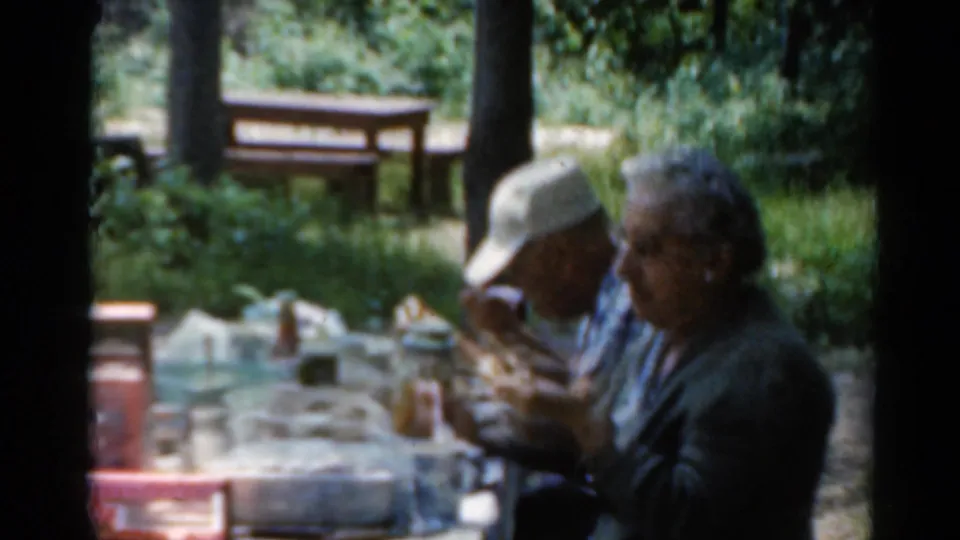 Two older adults, one in a white cap and patterned shirt, sit closely at a picnic table with food and drink items in Elkhart, Illinois.