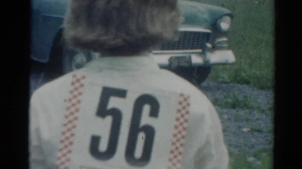 A person seen from behind wears a jacket marked 56 near a mid-century car, adding race-day context to the 1964 Elkhart home-movie scene.