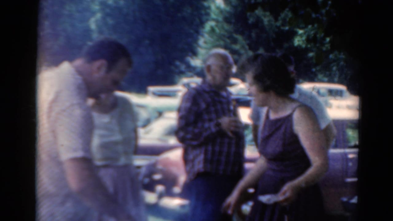 1964 Elkhart, Illinois home-movie frame showing adults gathered outdoors near vintage cars and trees.