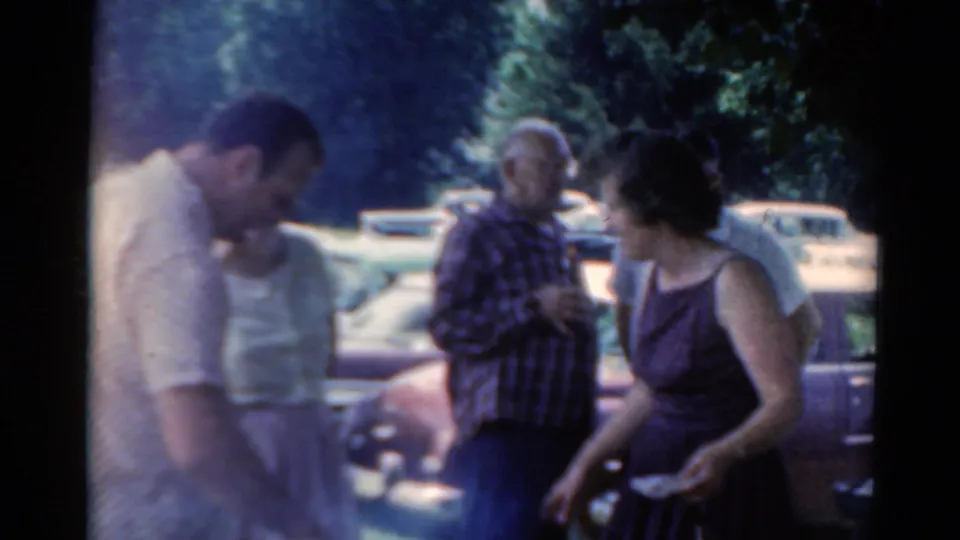 1964 Elkhart, Illinois home-movie frame showing adults gathered outdoors near vintage cars and trees.