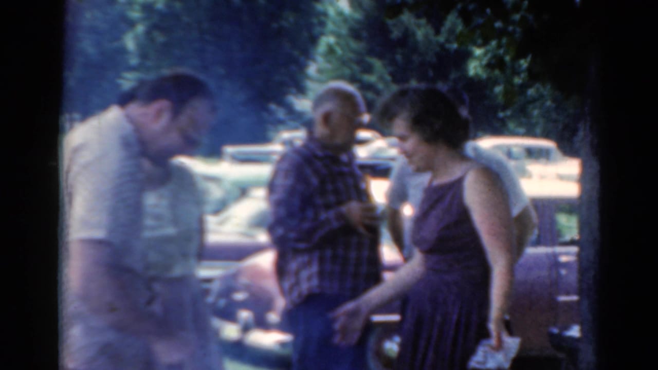 Adults in a 1964 Elkhart outdoor gathering stand near classic American cars with trees behind them.