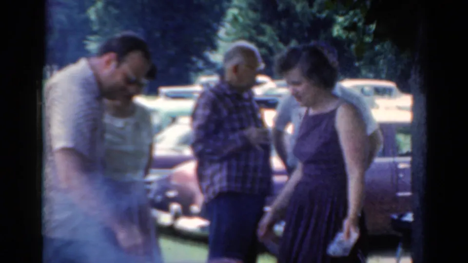 Home-movie frame from 1964 Elkhart showing adults moving and talking near parked vintage cars.