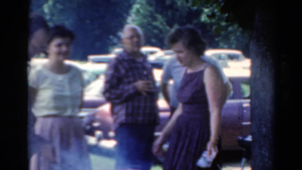 1964 Elkhart, Illinois frame of adults gathered by vintage cars, with a woman in a purple dress in the foreground.