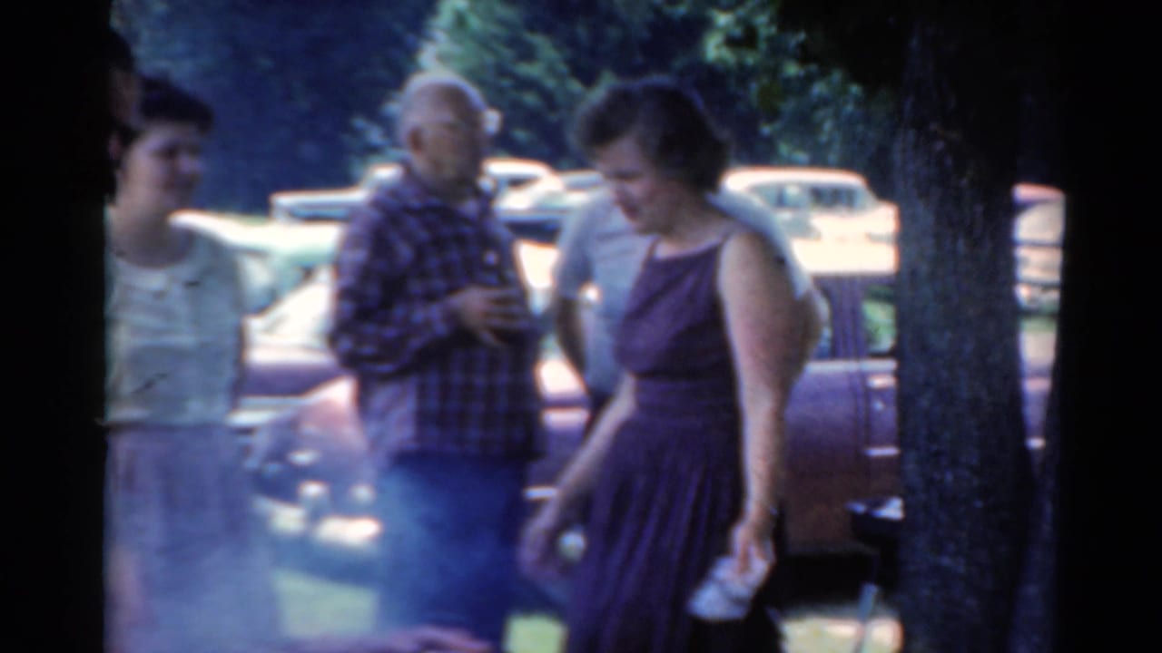 Three adults stand near a smoky outdoor grill with parked cars and trees behind them.