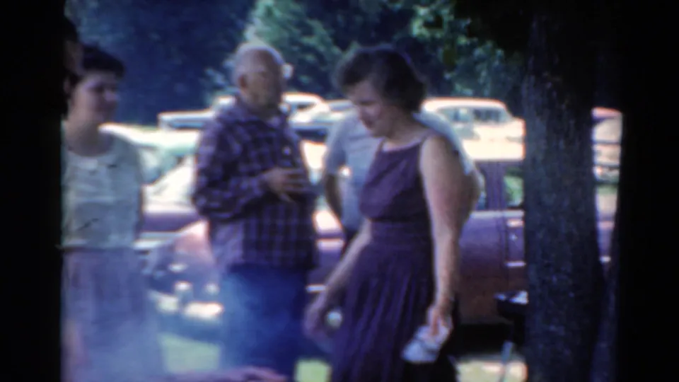 1964 Elkhart, Illinois home-movie frame showing adults gathered by a smoky grill, with trees and mid-century cars in the background.