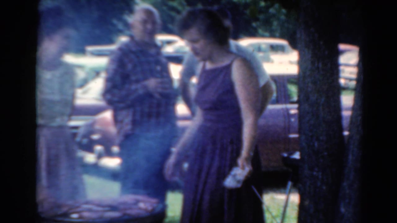 1964 Elkhart, Illinois frame of two adults by a smoky grill area, with trees and parked period cars behind them.