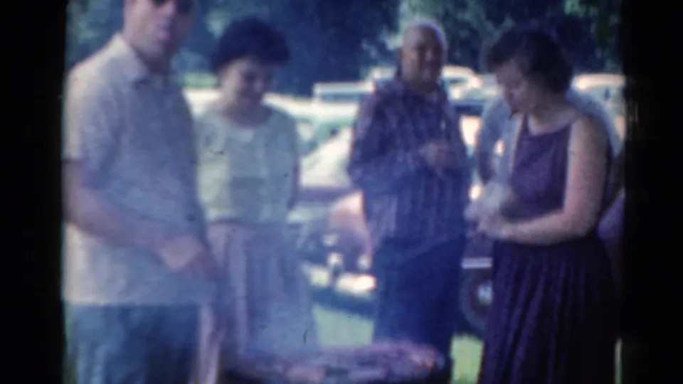 1964 Elkhart, Illinois home-movie frame showing four adults around a backyard grill, with smoke, trees, and parked cars visible.