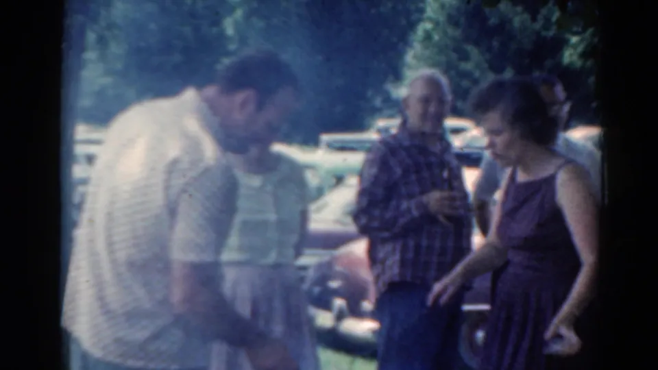 1964 Elkhart, Illinois frame showing a man tending or looking at a smoky grill while other adults stand near parked cars and trees.
