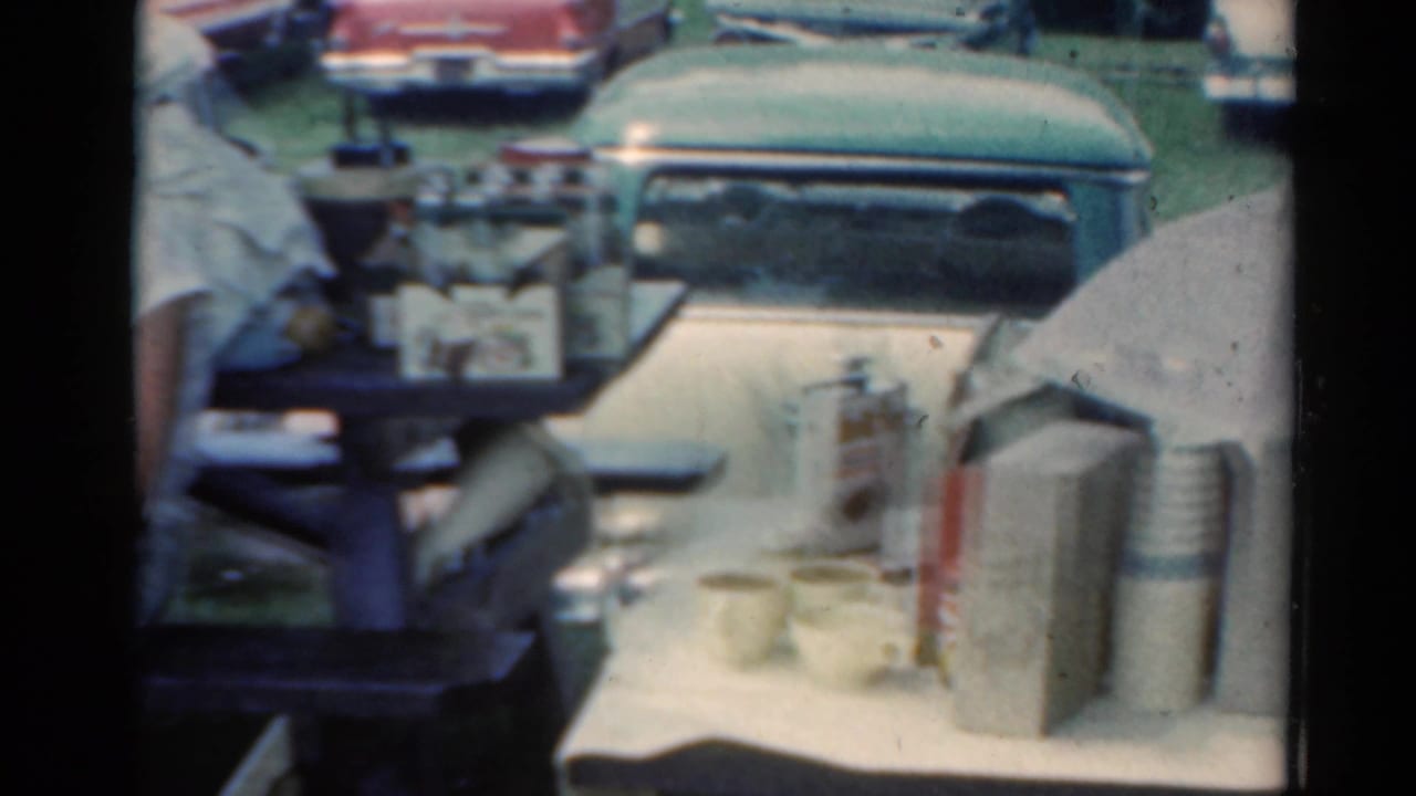 Picnic table with cups, thermos, umbrella, and parked cars behind it.