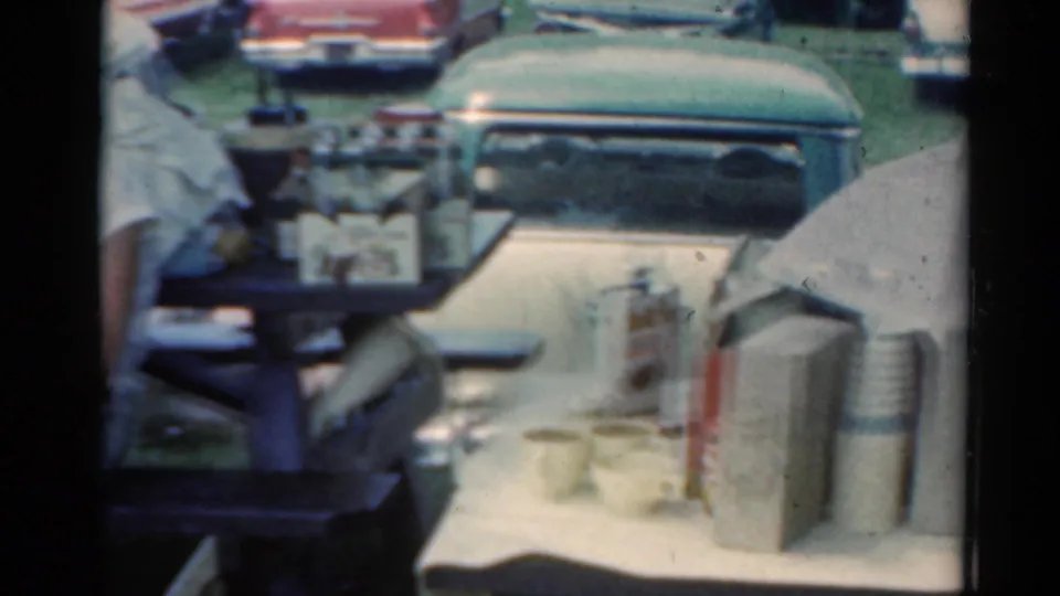 1964 Elkhart, Illinois home-movie frame showing a refreshment table with cups, a thermos, a paper umbrella, and classic cars behind it.