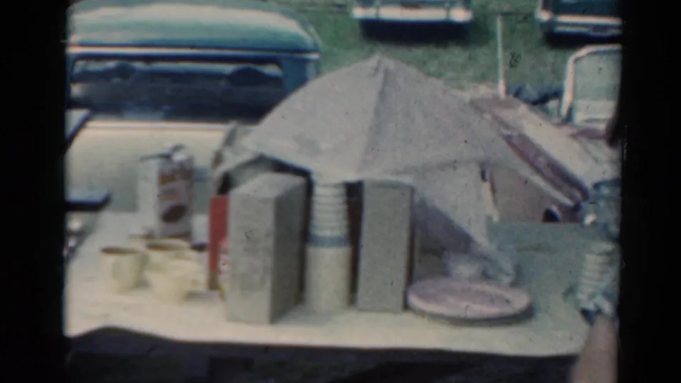 Outdoor Elkhart, Illinois scene with tabletop refreshments and classic cars visible behind, filmed in 1964 on 8mm stock.