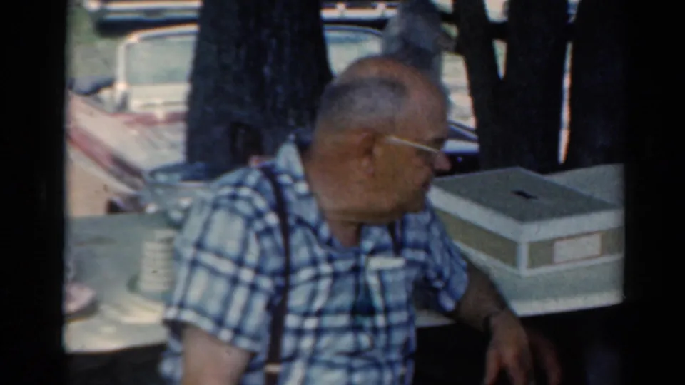 An older man in a blue plaid shirt sits at a picnic table during a 1964 Elkhart outdoor gathering, with cars and trees behind him.