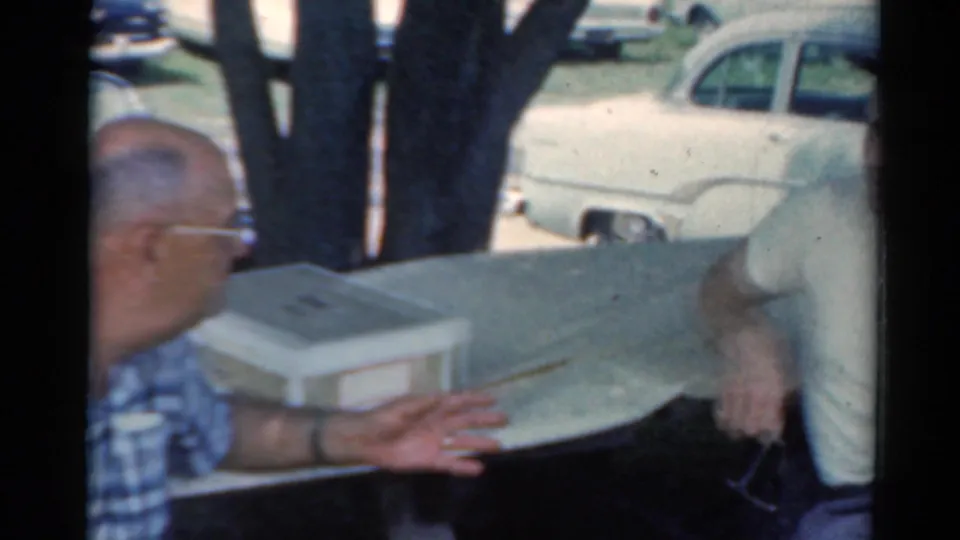 Two adult men appear in conversation at an outdoor table in Elkhart, Illinois, with cups, trees, and classic cars visible in the 1964 home movie frame.