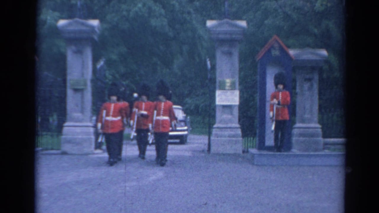 Uniformed men in red coats march near a stone gate while a sentry stands by a guard box.