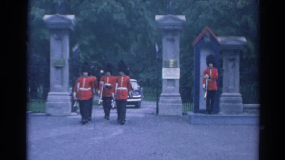 1964 8mm frame of red-coated uniformed men with rifles moving near a stone gate as a sentry stands at the entrance.