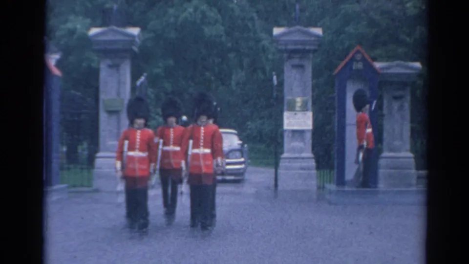 A small group of uniformed men in red coats walks toward a stone gate in supported 1964 8mm footage.