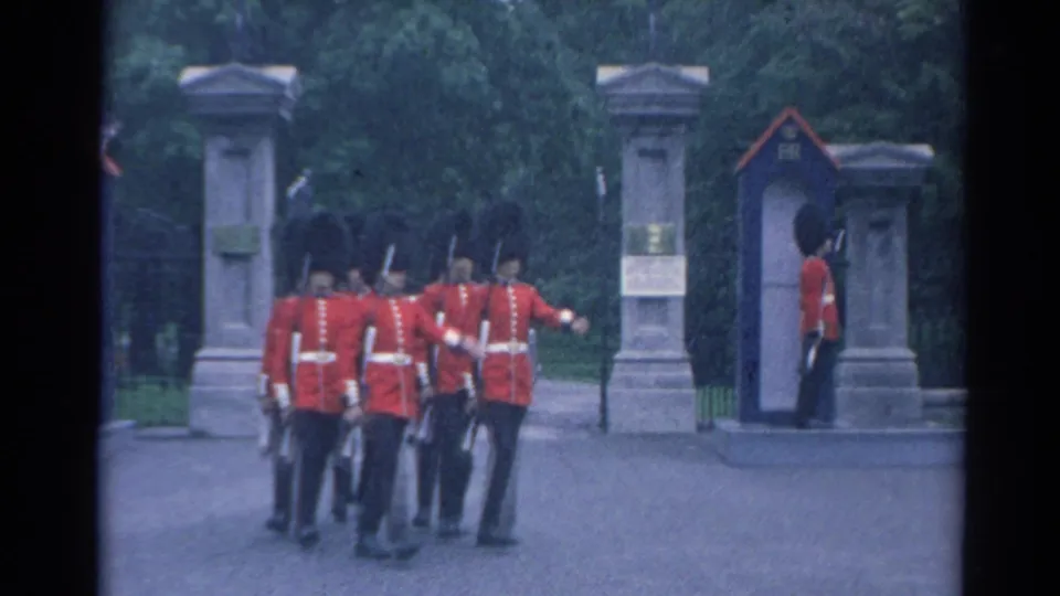 Red-coated uniformed men pause near a gated entrance while an armed sentry stands beside the guard box.