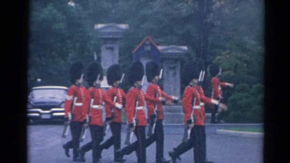 British-style guards march across the frame in red tunics and bearskin hats, with a dark car and gateposts in the background.