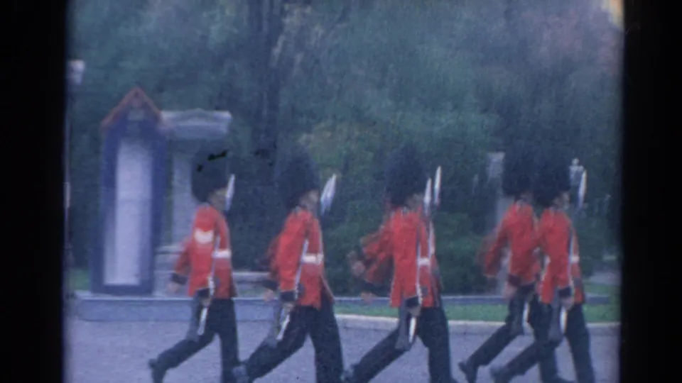 Soft 8mm frame of guards in red ceremonial uniforms moving past a gate structure and trees, with motion blur from the march.