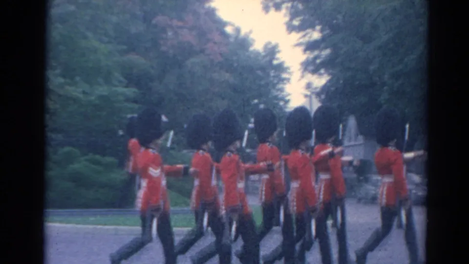A group of British-style guards in red tunics marches away along a tree-lined road in the 1964 home-movie footage.