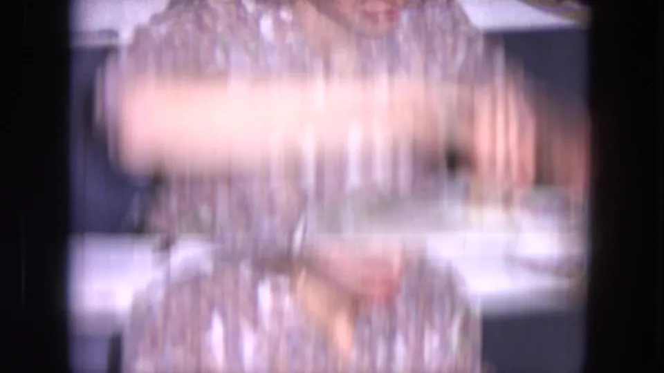 Motion-blurred 1964 Evanston home-movie frame of a woman in a patterned blouse near a kitchen table or counter.