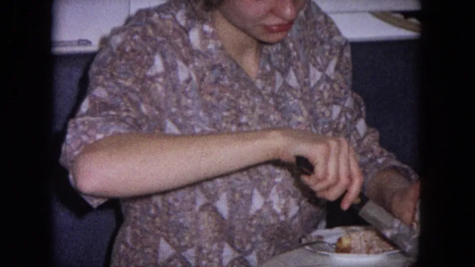 In a 1964 Evanston kitchen interior, a woman in a patterned blouse carefully cuts into a white iced cake.