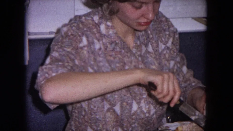 A woman in a patterned blouse looks down as she portions a white iced cake in a 1964 Evanston home kitchen.