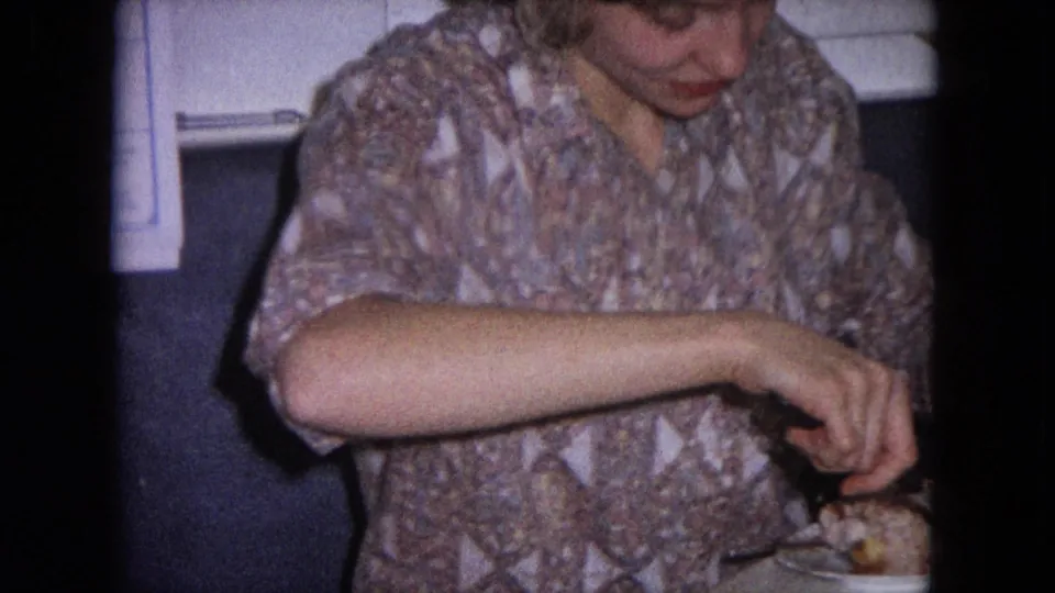 The woman looks down toward the plate as she carves or serves meat in the close-framed 8mm kitchen scene.