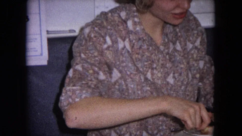 The woman remains seated at the table, her arm extended as she continues preparing food in the 1964 home-movie scene.