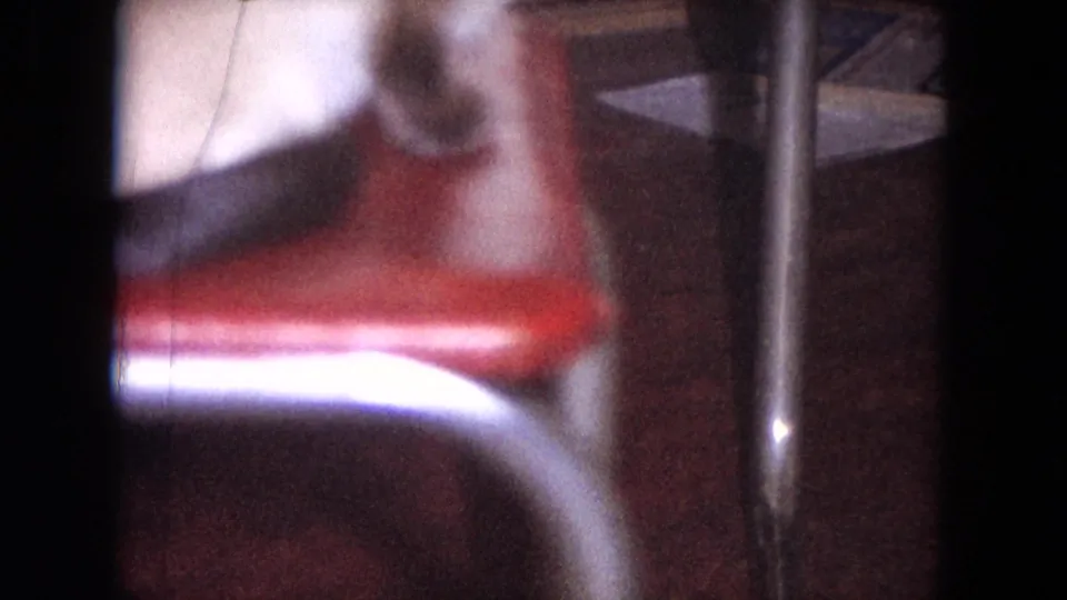 The camera drops toward a blurred red chair and table leg while the woman is only partly visible at the top of the home-movie frame.