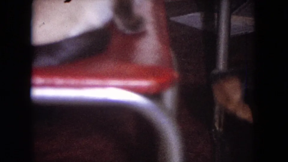 A soft, off-center home-movie frame shows a red chair, vertical table leg, and only a partial glimpse of the seated woman.
