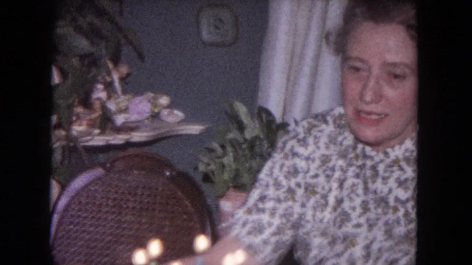 Seated woman in a floral blouse inside a 1964 Evanston home, with plants, curtains, wall decor, and a woven chair visible.
