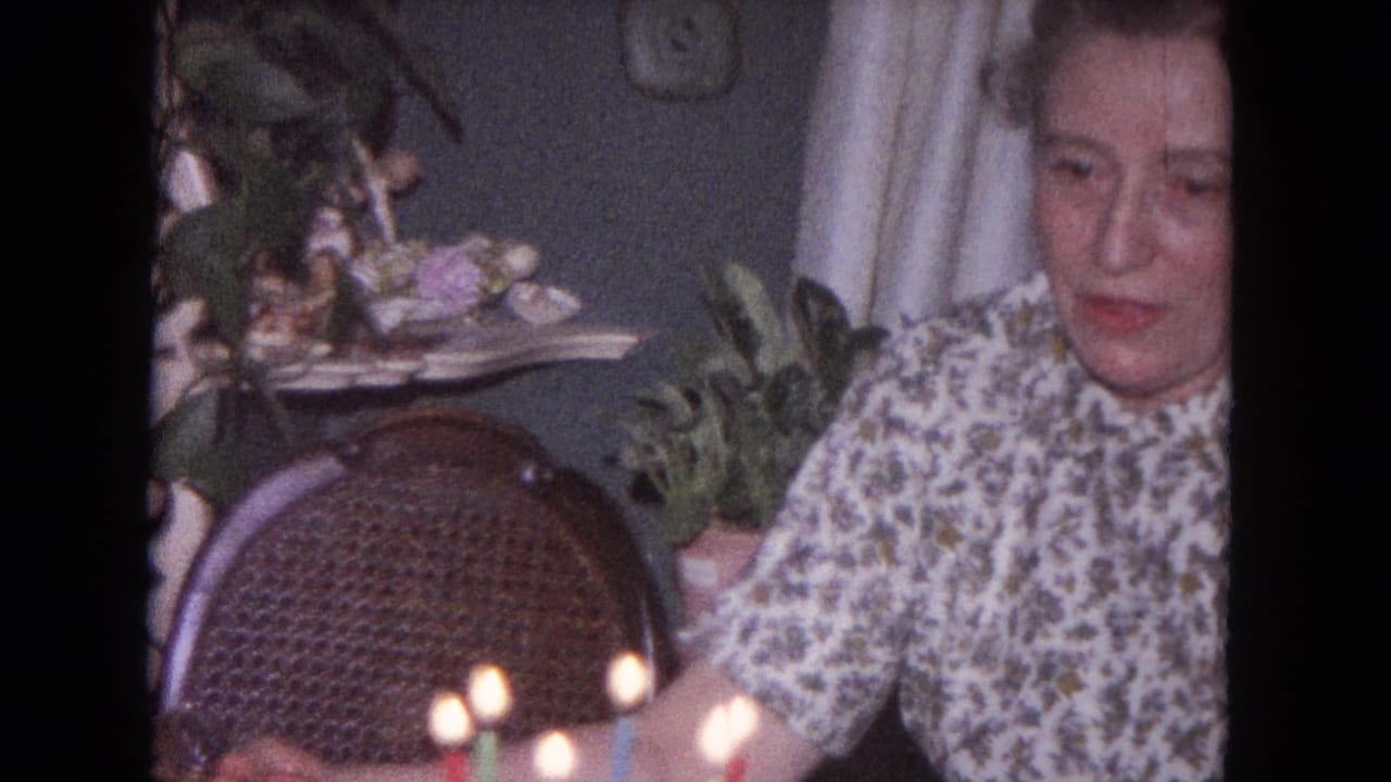 Woman seated indoors near lit birthday candles, with a wicker chair and houseplants behind her.