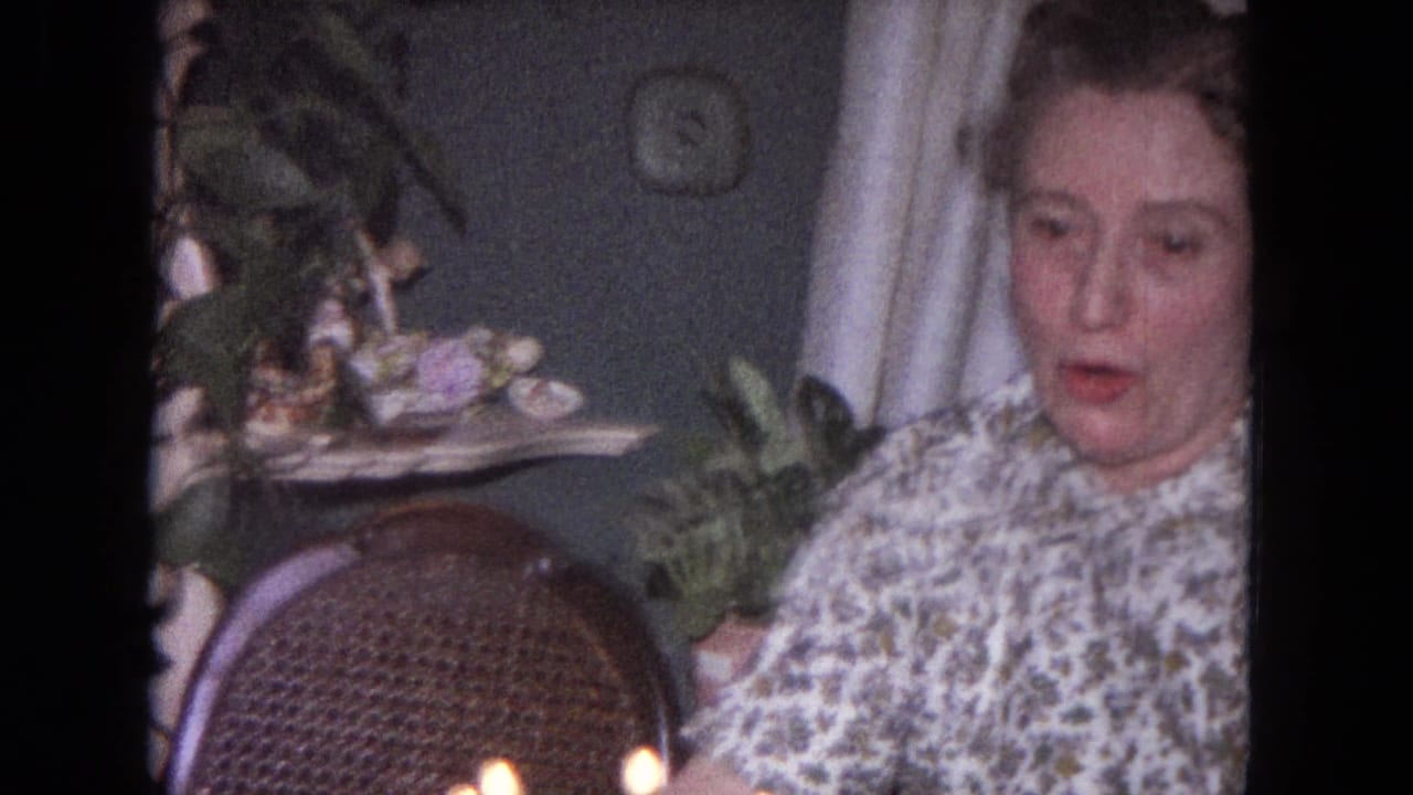 The woman remains seated beside birthday candles in a 1964 Evanston domestic setting, with houseplants and a wicker chair behind her.