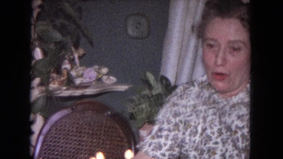 The woman remains seated beside birthday candles in a 1964 Evanston domestic setting, with houseplants and a wicker chair behind her.