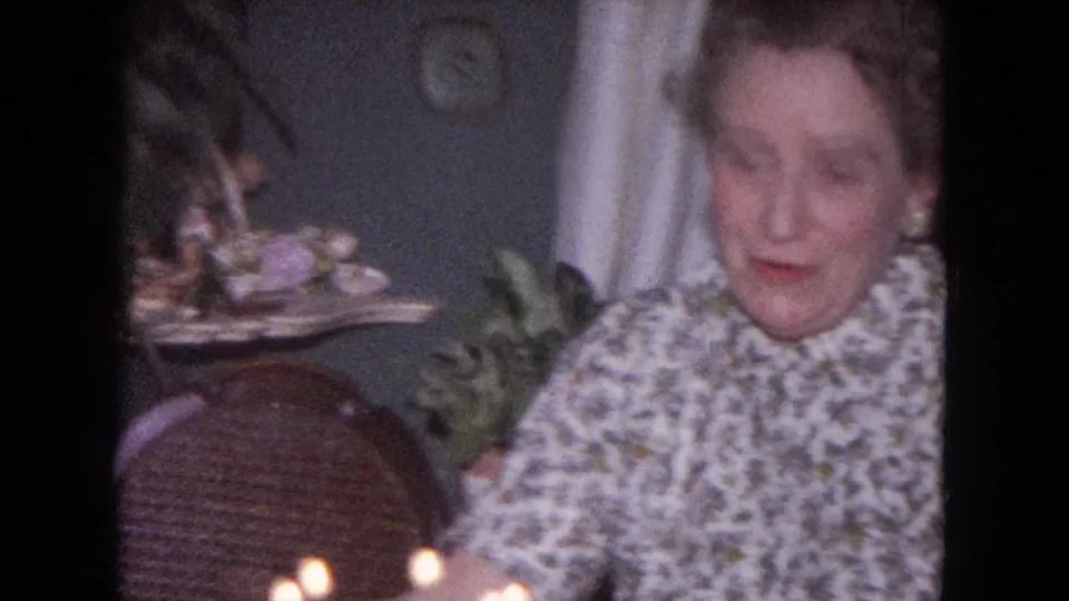 A softly blurred 8mm frame shows the woman moving at the table during the Evanston birthday scene, with candles in the foreground.