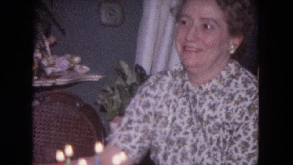 The woman smiles toward someone off camera in this 1964 Evanston home-movie frame, with birthday candles glowing nearby.